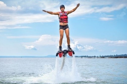 Person wearing helmet and vest flyboarding above water on a clear day.