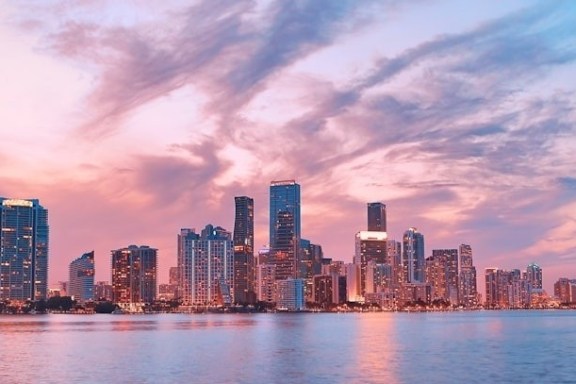 City skyline at sunset with colorful sky and water in foreground.